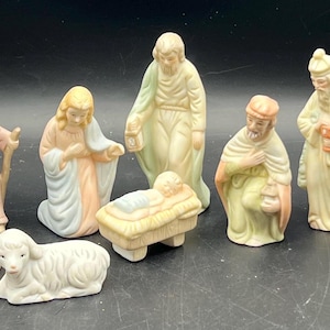 May include: A set of ten porcelain nativity figurines featuring figures in pastel tones of pink, blue, green, and cream. The collection includes a central infant in a manger, standing and kneeling figures, a donkey, and a lamb, all with matte finishes.