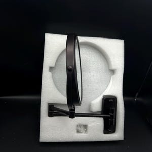 May include: A round, black, wall-mounted mirror with an adjustable arm. The mirror is set in a black frame and is packaged in white foam. The mirror is designed for bathroom or vanity use.