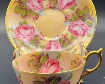 Può includere: Un delicato set da tè composto da tazza e piattino con un motivo floreale. Il set presenta uno sfondo giallo pallido con rose rosa e foglie verdi. La tazza e il piattino hanno un bordo dorato.