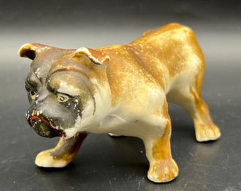 May include: A ceramic bulldog figurine in a standing pose. The dog has a brown and tan coat with a black face and red accents on the mouth. The figurine has a vintage appearance.