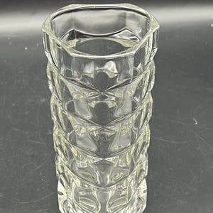 May include: A clear, faceted glass vase with a geometric design. The vase has a hexagonal top and base, with a series of diamond-shaped facets along its cylindrical body. The vase is empty and reflects light.