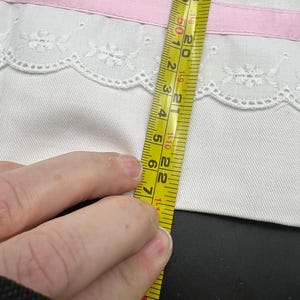 May include: White fabric with a pink ribbon and lace trim. The fabric is being measured with a yellow tape measure. The tape measure shows a measurement of approximately 7 inches.