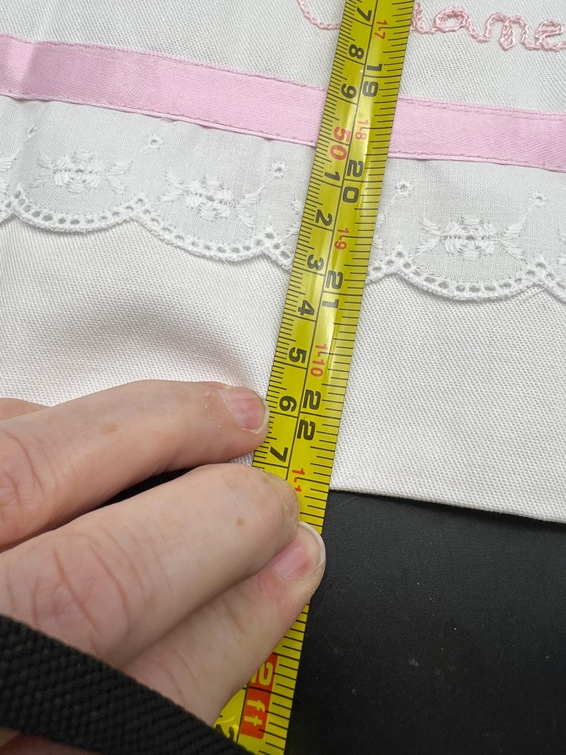 May include: White fabric with a pink ribbon and lace trim. The fabric is being measured with a yellow tape measure. The tape measure shows a measurement of approximately 7 inches.