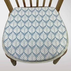 May include: A round chair cushion featuring a light blue backdrop and a repeating white leaf design. The cushion is placed on a wooden chair with a light brown finish. The cushion has a cream-coloured border.