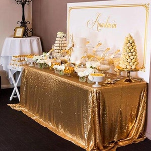 Könnte beinhalten: Eine goldene Sequinentischdecke, die über einen Tisch mit einem Dessertbuffet gelegt wird. Der Tisch ist mit weißen Blumen, Glasbehältern gefüllt mit Süßigkeiten und einer weißen Torte dekoriert. Ein Schild hinter dem Tisch lautet "Anastasia".