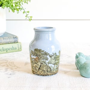 May include: A small, light blue ceramic vase with a hunting scene design. The scene features a man on horseback, a fox, and other animals in a forest setting.