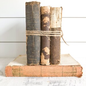 May include: Three vintage books with brown leather covers and worn spines. The books are tied together with twine and stacked on top of a larger book with a pink cover.