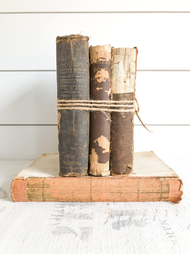 May include: Three vintage books with brown leather covers and worn spines. The books are tied together with twine and stacked on top of a larger book with a pink cover.