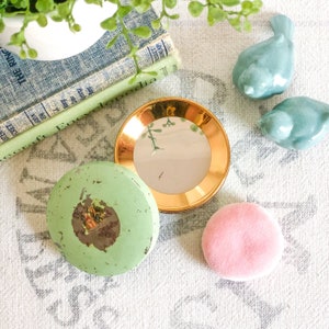 Puede incluir: Un conjunto de tocador vintage verde y dorado con un espejo redondo, una borla de polvo y un frasco con tapa. El conjunto está sobre un mantel de lino blanco con dos figuritas de pájaros azules.