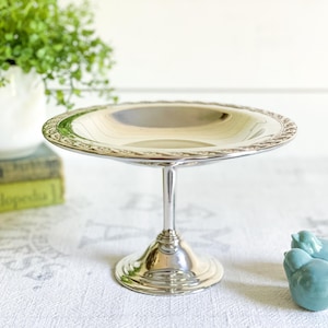 May include: A silver-colored, stemmed serving dish with a decorative rim. The dish is round and sits atop a slender stem and a flared base. A small, light blue ceramic bird figurine is to the right of the dish.