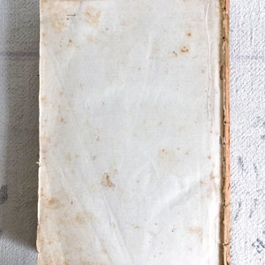 May include: A vintage book with a white cover and a brown spine. The cover has several brown stains.