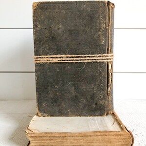 May include: Two antique books stacked on top of each other. The top book is bound in dark brown leather and has a twine string tied around it. The bottom book is bound in a lighter brown paper and has a worn, aged appearance.