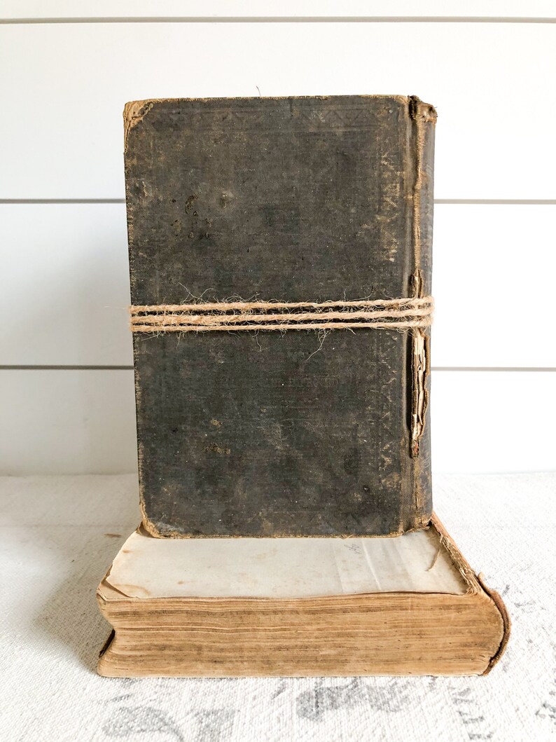 May include: Two antique books stacked on top of each other. The top book is bound in dark brown leather and has a twine string tied around it. The bottom book is bound in a lighter brown paper and has a worn, aged appearance.