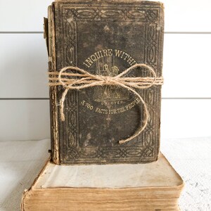 May include: A vintage, brown leather-bound book titled "Inquire Within, Or, Over 3,700 Facts for the People" with a twine bow tied around it. The book is sitting on top of another, older book.