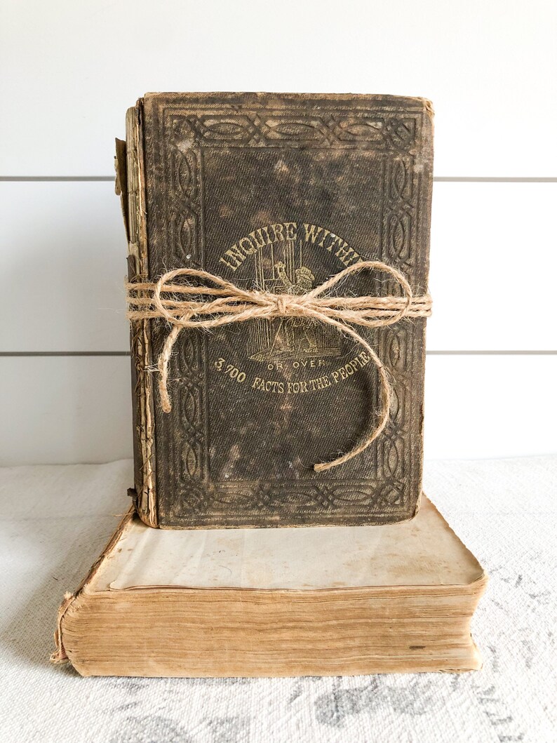 May include: A vintage, brown leather-bound book titled "Inquire Within, Or, Over 3,700 Facts for the People" with a twine bow tied around it. The book is sitting on top of another, older book.