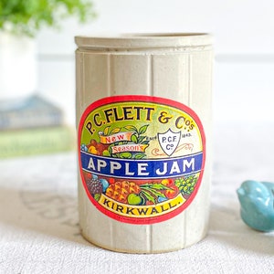 Puede incluir: Un tarro de gres vintage con una etiqueta colorida que dice "P.C. Flett & Co's. Est. 1943. New Season's Apple Jam. Kirkwall".