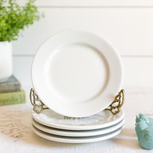 May include: A set of four white ceramic plates stacked on a gold metal stand. The plates are plain and simple, with a smooth, white finish.