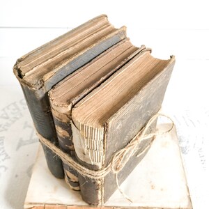 May include: A stack of three vintage hardcover books tied together with twine. The books are worn and have a rustic look. The books are sitting on a white surface.
