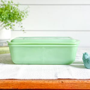 Peut inclure: Un plat rectangulaire en verre vert clair avec un couvercle assorti. Le plat est posé sur un tissu blanc sur une surface en bois. Une petite figurine d'oiseau bleu se trouve à droite du plat.