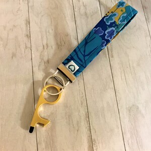 May include: A gold-colored key chain with a blue and yellow floral pattern fabric strap. The key chain has a loop and a tool with a bottle opener and a hook.