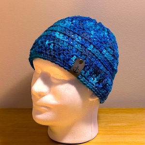 May include: Blue and teal crocheted beanie with an anchor-shaped leather tag. The beanie is on a white mannequin head.