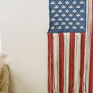 Puede incluir: Un tapiz de macram&eacute; que representa la bandera estadounidense. La esquina superior izquierda presenta un campo azul con nudos blancos en forma de estrella. Rayas verticales rojas y blancas forman la parte inferior, con flecos en la parte inferior. Dise&ntilde;ado para colgar.