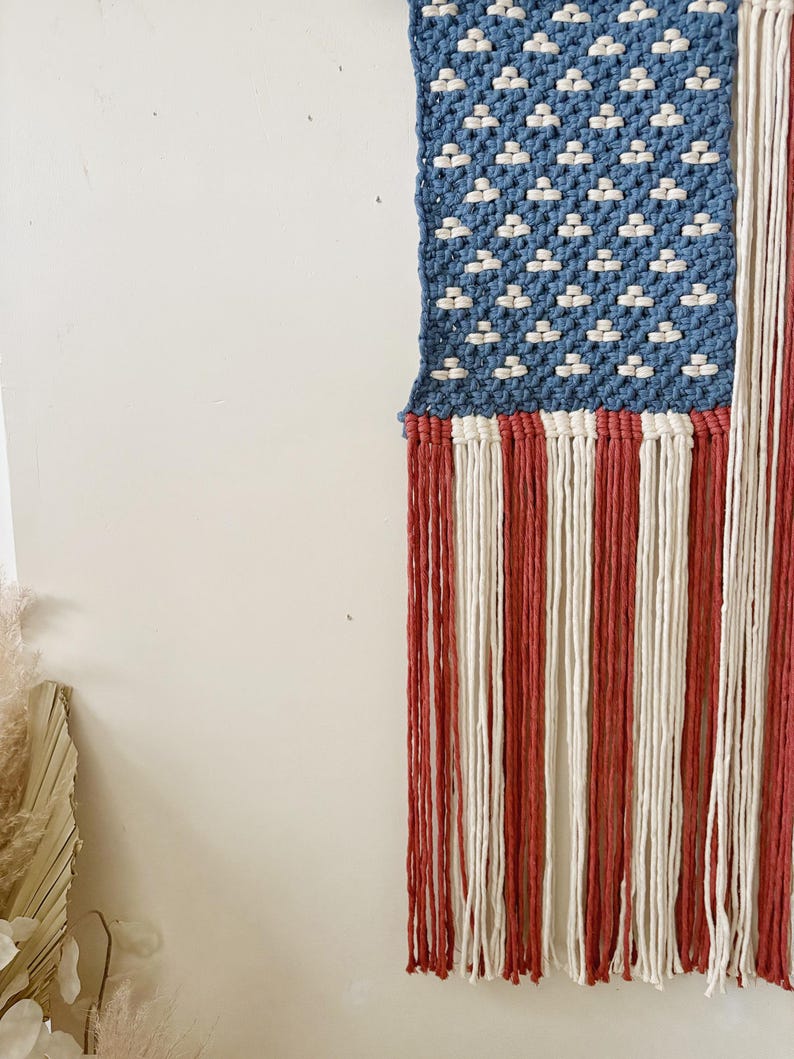 Puede incluir: Un tapiz de macram&eacute; que representa la bandera estadounidense. La esquina superior izquierda presenta un campo azul con nudos blancos en forma de estrella. Rayas verticales rojas y blancas forman la parte inferior, con flecos en la parte inferior. Dise&ntilde;ado para colgar.