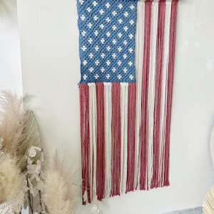 Puede incluir: Un tapiz de macram&eacute; que representa la bandera estadounidense. La secci&oacute;n superior es azul con patrones blancos en forma de estrella, y la secci&oacute;n inferior presenta rayas verticales rojas y blancas. La bandera est&aacute; suspendida de un palillo de madera.