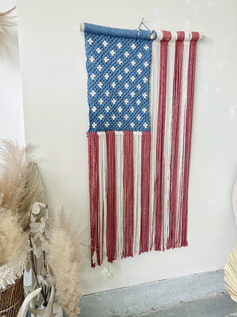 Puede incluir: Un tapiz de macram&eacute; que representa la bandera estadounidense. La secci&oacute;n superior es azul con patrones blancos en forma de estrella, y la secci&oacute;n inferior presenta rayas verticales rojas y blancas. La bandera est&aacute; suspendida de un palillo de madera.