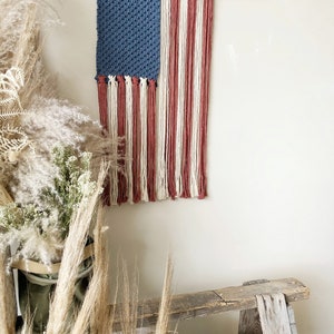 May include: A handmade macrame wall hanging in the shape of the American flag. The flag is made with blue, white, and red yarn and is hung on a wooden branch.