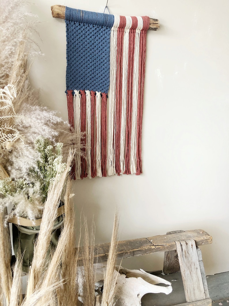May include: A handmade macrame wall hanging in the shape of the American flag. The flag is made with blue, white, and red yarn and is hung on a wooden branch.