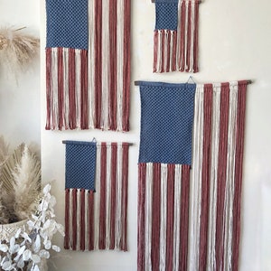 May include: Four woven wall hangings in the shape of the American flag. The flags are made with blue, white, and red yarn. The flags are different sizes and are hanging on a white wall.