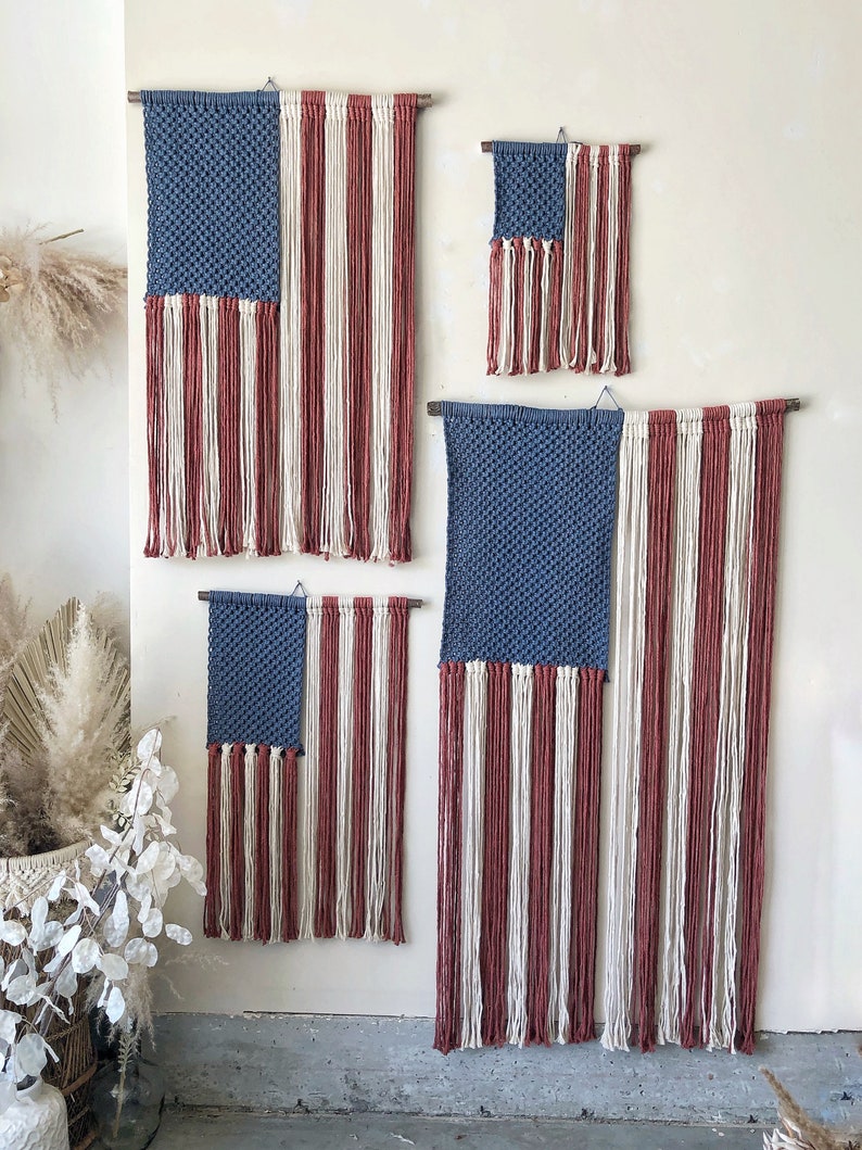 May include: Four woven wall hangings in the shape of the American flag. The flags are made with blue, white, and red yarn. The flags are different sizes and are hanging on a white wall.