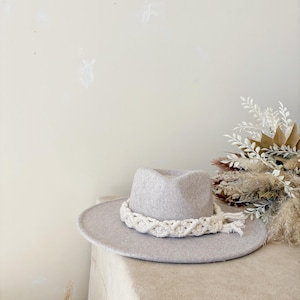 May include: A light gray fedora hat with a wide brim and a cream-colored, braided band. The hat is displayed on a neutral-toned surface, with dried floral arrangements in the background. This stylish hat is a fashionable accessory.