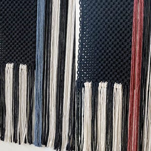 May include: Two macrame wall hangings with black, white, and blue yarn. The left hanging has a blue and white striped pattern, while the right hanging has a red and white striped pattern.