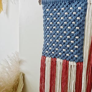 Puede incluir: Un tapiz de macram&eacute; con el dise&ntilde;o de la bandera estadounidense. La secci&oacute;n superior es azul con nudos blancos, y la parte inferior tiene flecos rojos y blancos. El tapiz est&aacute; montado sobre un trozo de madera.