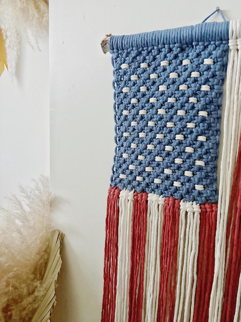 Puede incluir: Un tapiz de macram&eacute; con el dise&ntilde;o de la bandera estadounidense. La secci&oacute;n superior es azul con nudos blancos, y la parte inferior tiene flecos rojos y blancos. El tapiz est&aacute; montado sobre un trozo de madera.