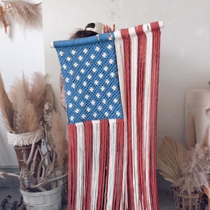 Puede incluir: Colgante de pared de macram&eacute; que representa la bandera estadounidense. El campo azul presenta estrellas blancas, mientras que las rayas alternan entre rojo y blanco. La bandera est&aacute; suspendida de un list&oacute;n de madera, una decoraci&oacute;n patri&oacute;tica.