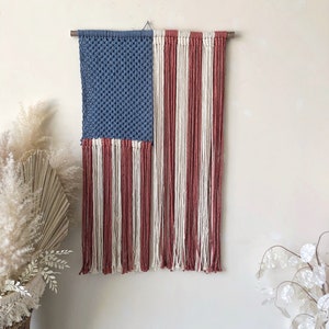 May include: A handmade macrame wall hanging in the shape of the American flag. The flag is made with blue, white, and red yarn. The flag is hanging on a wooden dowel rod.