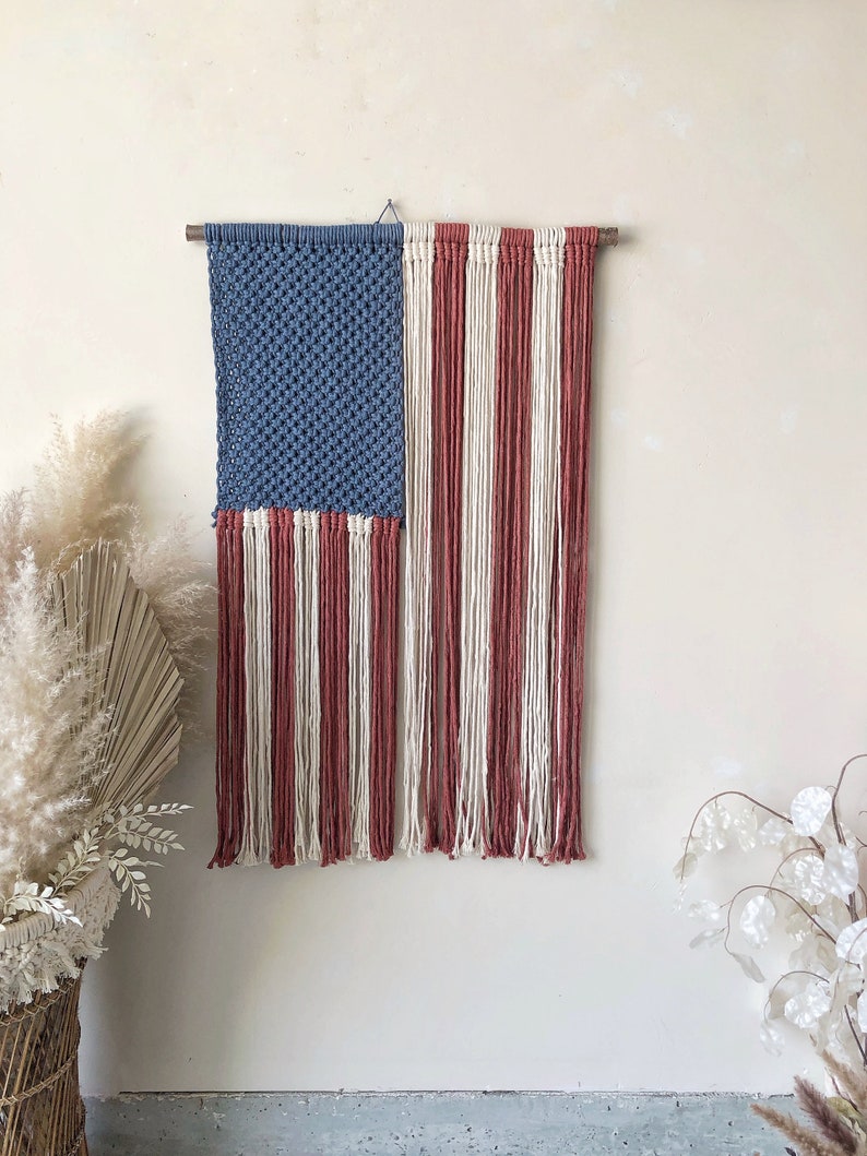 May include: A handmade macrame wall hanging in the shape of the American flag. The flag is made with blue, white, and red yarn. The flag is hanging on a wooden dowel rod.