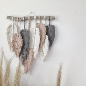 May include: A wall hanging made of macrame feathers in shades of white, gray, and pink. The feathers are attached to a natural wood branch.
