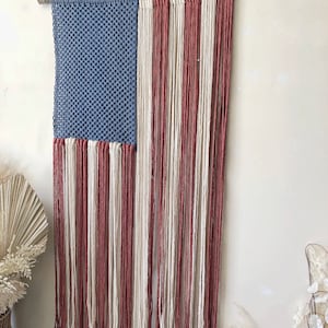 May include: A handmade macrame wall hanging in the shape of the American flag. The flag is made with blue, white, and red yarn. The flag is hanging from a wooden dowel rod.