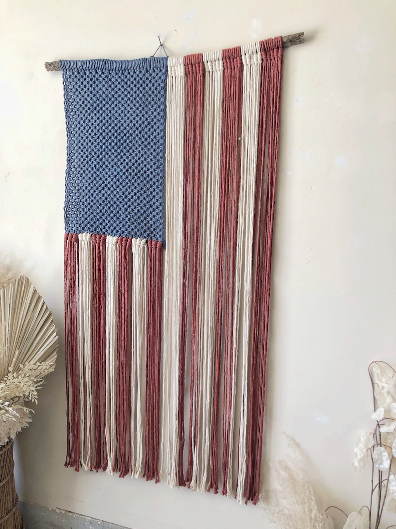 May include: A handmade macrame wall hanging in the shape of the American flag. The flag is made with blue, white, and red yarn. The flag is hanging from a wooden dowel rod.