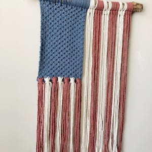 May include: A woven wall hanging in the shape of the American flag. The flag is made of blue, red, and white yarn and is attached to a piece of driftwood.