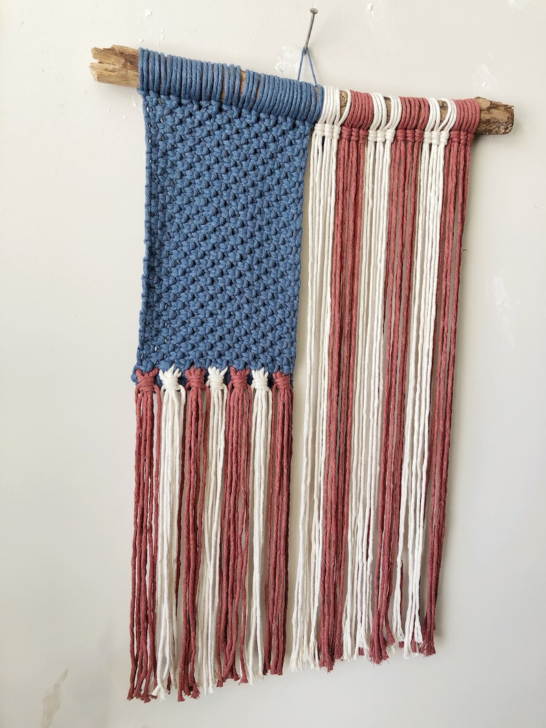 May include: A woven wall hanging in the shape of the American flag. The flag is made of blue, red, and white yarn and is attached to a piece of driftwood.