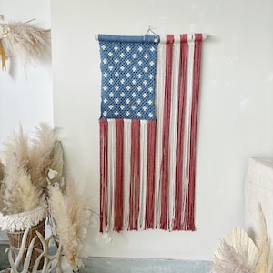 Puede incluir: Un tapiz de macram&eacute; con la forma de la bandera estadounidense. La secci&oacute;n superior es azul con patrones blancos en forma de estrella, y la inferior tiene rayas verticales rojas y blancas. Cuelga de una barra de madera.