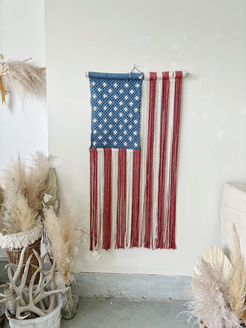 Puede incluir: Un tapiz de macram&eacute; con la forma de la bandera estadounidense. La secci&oacute;n superior es azul con patrones blancos en forma de estrella, y la inferior tiene rayas verticales rojas y blancas. Cuelga de una barra de madera.