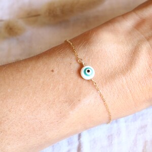 Peut inclure: Un bracelet chaîne de couleur or avec un charm central représentant un œil. Le charm a une base blanche avec un détail d'œil bleu et noir. Le bracelet est délicat et porté sur un poignet.