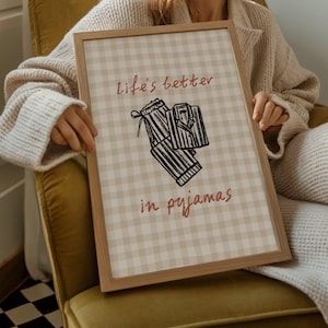 Pu&ograve; includere: Una stampa incorniciata con uno sfondo a motivo gingham marrone chiaro. La stampa presenta il testo "life's better in pyjamas" in un colore marrone rossiccio, insieme a un'illustrazione in bianco e nero di un pigiama. La cornice &egrave; in legno chiaro.