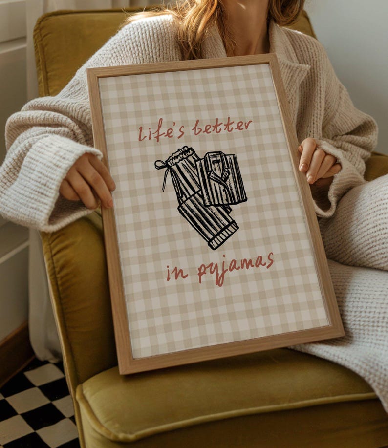 Pu&ograve; includere: Una stampa incorniciata con uno sfondo a motivo gingham marrone chiaro. La stampa presenta il testo "life's better in pyjamas" in un colore marrone rossiccio, insieme a un'illustrazione in bianco e nero di un pigiama. La cornice &egrave; in legno chiaro.